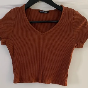 Brun ribbad croppad topp från SHEIN - Säljer en brun croppad topp från SHEIN i storlek S. Toppen har ribbad struktur, korta ärmar och en snygg v-ringad hals. Perfekt för dig som gillar enkel och trendig stil. Materialet är mjukt och stretchigt, vilket gör den skön att bära.