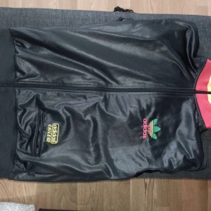 Adidas Chile 62 svart jacka - Svart Adidas Chile 62 jacka i syntetmaterial med glansig finish. Hög krage med gröna, gula och röda ränder. Broderad Adidas-logga på bröstet och gul brodyr på fickan. Dragkedja framtill och ficka med dragkedja. Perfekt för dig som gillar retrostil.