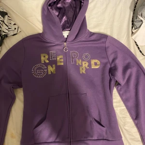 Lila hoodie från Freejoy med bling - Lila hoodie från Freejoy med glansigt innerfoder i huvan. Framsidan har tryck i guld och strassdetaljer. Dragkedja framtill och två fickor. Perfekt för dig som vill sticka ut med lite extra bling och färg.