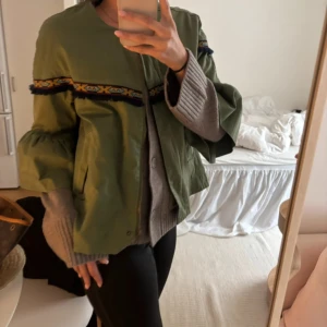 Grön overshirt med volangärmar - Säljer en grön overshirt med coola volangärmar och ett broderat band i blått, gult och rött över bröstet och ryggen. Jackan har en loose passform och är tillverkad i ett lätt syntetmaterial. Perfekt för dig som vill sticka ut med detaljer och färg.