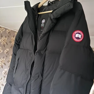 Svart pufferjacka från Canada Goose armstrong hoody - Svart pufferjacka från Canada Goose med ikonisk röd och vit logotyp på ärmen. Jackan har huva, dragkedja och stora fickor framtill. Fylld med dun för extra värme och har en loose passform. Perfekt för kalla vinterdagar och har en stilren look.