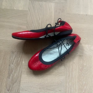 Röda Replay ballerinaskor med snörning - Snygga röda ballerinaskor från Replay med svarta detaljer och snörning över foten. Skorna har rund tå, svarta sulor med Replay-logga undertill och är tillverkade i glansigt syntetmaterial med dekorativa sömmar och små perforeringar framtill.