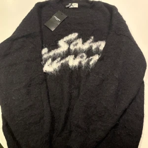 Svart stickad Saint Laurent tröja - Mjuk och fluffig svart stickad tröja från Saint Laurent med vit logga framtill. Tröjan har lång ärm och klassisk rund halsringning. Perfekt för dig som vill ha en lyxig och stilren statement piece i garderoben.