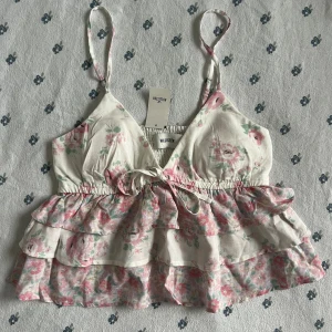 Blommig babydoll volangtop från Hollister - Supersöt blommig babydoll topp från Hollister med volanger och smala axelband. Toppen är vit med rosa och gröna blommor, har v-ringning, knyt framtill och uttagbara inlägg. Perfekt för sommarens alla tillfällen!