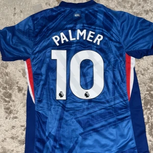 Chelsea FC Palmer 10 Nike fotbollströja M - Chelsea FC matchtröja med Palmer 10-tryck på ryggen. Tröjan är blå med vita och röda detaljer, har korta ärmar och är tillverkad i Dri-FIT-material som andas. Snyggt geometriskt mönster framtill och officiellt klubbmärke på bröstet.