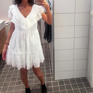 Vit volangklänning med v-ringning - Supersöt vit kortklänning med v-ringning och volangdetaljer både vid axlar och kjol. Klänningen är ärmlös med lätt transparent tyg och subtilt blommönster. En av mina studentklänningar, använd endast en gång på studentdagen, ser ut som helt ny. Pris kan diskuteras!