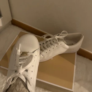 Vita sneakers från Michael Kors - Snygga vita sneakers från Michael Kors i skinn med klassisk rund tå och platt sula. Skorna har vita snören och guldfärgad MK-detalj framtill.  Använda endast en gång!!