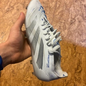 Adidas Predator fotbollsskor vita/blå - Säljer ett par Adidas Predator fotbollsskor i vitt med blå detaljer och gråa ränder. Skorna har snörning, platt sula med dobbar och är tillverkade i syntetmaterial. Perfekta för dig som vill sticka ut på planen med stilren design och grym känsla på bollen. Skorna är i nyskick