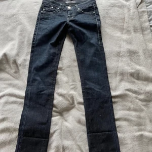 Rock & Republic mörka jeans med brodyr - Snygga mörkblå jeans från Rock & Republic med raka ben och coola broderade detaljer på bakfickorna. Klassisk femficksmodell med kontrastsömmar och silverfärgade nitar. Perfekta för dig som gillar en stilren men ändå unik look.