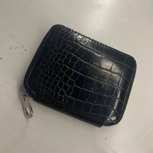 Svart plånbok i croco-mönstrat skinn - Snygg svart plånbok i croco-mönstrat skinn med dragkedja runtom. Plånboken har flera fack för kort och mynt, samt en silverfärgad dragkedja och metallhake. Perfekt storlek för att ha med sig överallt.