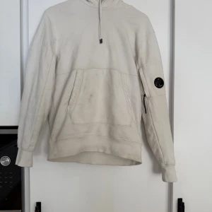 Vit hoodie från C.P. Company - Säljer en stilren vit hoodie från C.P. Company med klassisk huva och snörning. Tröjan har en stor magficka och en ikonisk svart linsdetalj på ärmen. Perfekt för dig som gillar streetwear och vill ha något bekvämt och snyggt.