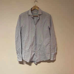 Randig skjorta från Tiger of Sweden - Säljer en slim fit skjorta från Tiger of Sweden i ljusblått och vitt med klassiskt randigt mönster. Skjortan har långa ärmar, knappar framtill och är tillverkad i bomull. Perfekt för dig som gillar stilrena och fräscha plagg.