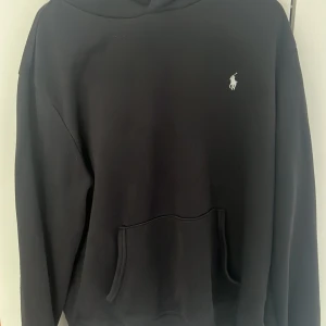 Svart hoodie från Polo Ralph Lauren - Svart hoodie från Polo Ralph Lauren med klassisk vit logga på bröstet. Tröjan har huva, känguruficka och långa ärmar. Perfekt för en chill och stilren look. Tillverkad i mjukt bomullsmaterial.