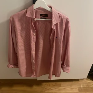 Randig skjorta från Morris, röd/vit - Säljer en klassisk röd- och vitrandig skjorta från Morris med blå broderad logga på bröstet. Skjortan har lång ärm, knappar framtill och är gjord i mjuk bomull. Perfekt för dig som gillar stilrena och tidlösa plagg. Andvänd två gånger