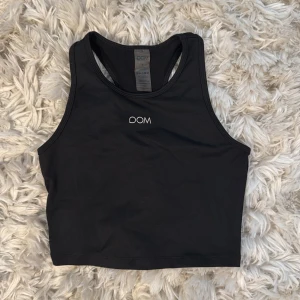Svart linnetopp från Drop of Mindfulness - Svart croppad linnetopp från Drop of Mindfulness med brottarrygg och vit logotyp framtill. Tillverkad i mjuk och stretchig polyesterblandning som sitter skönt mot kroppen. Perfekt för träning och aktiva dagar.aldrig använd