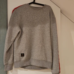 Grå sweatshirt Alpha Industries M - Snygg grå sweatshirt från Alpha Industries i bomull med röda detaljer längs axlar och ärmar där det står text. Tröjan har rund halsringning, långa ärmar och ribbade muddar. Perfekt för en avslappnad och cool stil.
