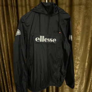 Svart vindjacka från Ellesse - Svart vindjacka från Ellesse med stor logga framtill och på ärmarna. Jackan har huva med snörning, dragkedja upptill och en stor ficka på magen. Tillverkad i lätt polyester, perfekt för blåsiga dagar. Snygg och sportig stil som sticker ut.