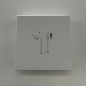 Apple AirPods (2nd Generation) - Apple AirPods (2nd gen) i originalförpackning med laddningsetui och laddkabel. Trådlösa hörlurar med smidig anslutning till iPhone och andra Apple-enheter. Produkten ser ut att vara i mycket gott skick utan synligt slitage.