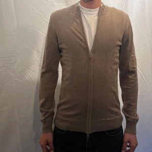 Ziptröja från Stenströms - Snygg beige zip tröja från Stenströms med hel dragkedja framtill och ribbad krage. Tillverkad i mjuk ull och har en klassisk, smal passform. Perfekt för lager-på-lager och funkar året runt. Enkel och stilren design som passar till det mesta. Modellen är 180cm.