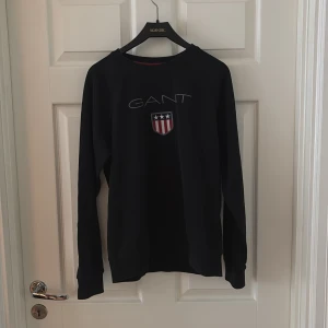 Svart långärmad tröja från GANT - Svart långärmad tröja från GANT med rund halsringning och broderad logga samt sköld på bröstet. Klassisk och stilren design som passar perfekt till jeans eller chinos. Mjuk och bekväm bomullskvalitet. Barnstorlek