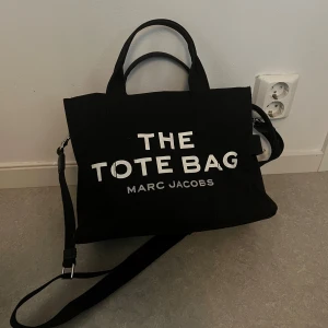 Svart tote bag från Marc Jacobs - Säljer en svart tote bag från Marc Jacobs med tydlig vit logga framtill. Väskan har både handtag och justerbar axelrem, samt dragkedja upptill. Tillverkad i slitstark bomullscanvas och rymmer allt du behöver till skolan eller stan. Köpt på Nelly så digitalt kvitto finns. Medium storlek 