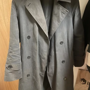 Grå trenchcoat med knappar - Stilren grå trenchcoat med dubbelknäppning och klassisk krage. Jackan har långa ärmar med knappar vid ärmslut och är perfekt för dig som gillar en tidlös look. Passar bra till lager-på-lager och har en rak, lite loose passform.