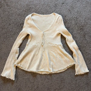 Beige ribbad blus med snörning - Superfin beige ribbad blus med v-ringning och snörning framtill. Långa ärmar och volangkant vid ärmslut och nederkant ger en söt touch. Perfekt för dig som gillar mjuka färger och sköna material.