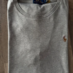 Grå t-shirt från Polo Ralph Lauren - Snygg grå t-shirt från Polo Ralph Lauren i bomull med klassisk rund hals och broderad logga på bröstet. Modellen är slim fit och passar perfekt till jeans eller shorts. Enkel och stilren design som funkar året runt.