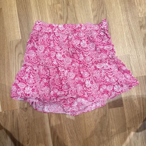 Rosa blommig kortkjol från Zara - Supersöt kortkjol från Zara i rosa och vitt med blommigt mönster. Kjolen har volangdetaljer och resår i midjan för en bekväm passform. Perfekt för varma dagar och ger en fräsch och somrig vibe.