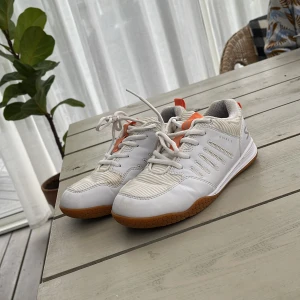 Vita Rival 2 innebandyskor med orange detaljer - Säljer ett par vita Rival 2 sneakers med snygga orange detaljer och bruna gummisulor med bikakemönster. Skorna har snörning och en sportig design med textil och syntetmaterial. Perfekta för dig som gillar stilrena och fräscha innebandyskor