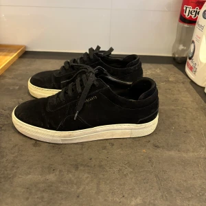 Svarta sneakers från Axel Arigato - Svarta sneakers från Axel Arigato i mocka med vit platt sula och klassisk snörning. Snygg minimalistisk design med diskret guldfärgad logga på sidan. Perfekta för dig som gillar stilrena och trendiga skor.