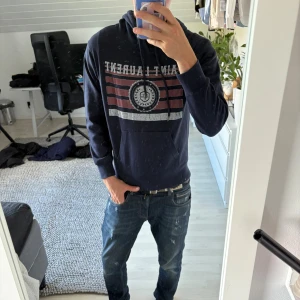 Mörkblå Saint Laurent hoodie  - Säljer en mörkblå hoodie från Saint Laurent med tryck framtill i vitt och rött. Tröjan har snörning i huvan, känguruficka och är tillverkad i mjuk bomull. Perfekt för dig som vill ha en stilren och exklusiv streetlook.