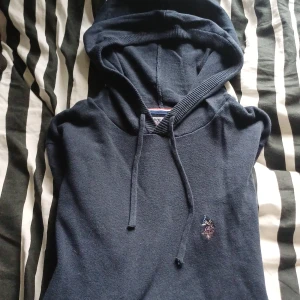 Mörkblå hoodie från U.S. Polo Assn. - Säljer en mörkblå hoodie från U.S. Polo Assn. med broderad logga på bröstet. Tröjan har huva med snörning och är tillverkad i ett stickat material som känns både mjukt och stilrent. Perfekt för dig som gillar klassisk och sportig stil.