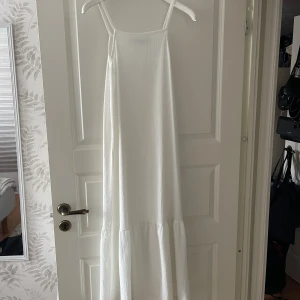 Vit långklänning med smala axelband - En stilren vit långklänning med smala axelband och lätt rynkad volang nedtill. Klänningen har en rak, luftig passform och är tillverkad i ett tunt, strukturerat tyg som ger en somrig känsla. Perfekt för varma dagar och enkel att styla med accessoarer.