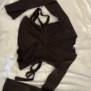 Svart blus från Nelly med knyt bak  - Svart blus från Nelly med knyt bak. Långa ärmar och v-ringning. Toppen har ribbad struktur och knytband i midjan som ger en snygg siluett. Perfekt att styla med jeans eller kjol för en trendig look.