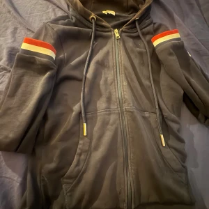 Svart Moncler hoodie med ränder - Svart hoodie från Moncler med dragkedja och huva. Snygga röda, vita och grå ränder på ärmarna och huvan ger en sportig vibe. Guldiga detaljer på snörningen och Moncler-logga på bröstet. Perfekt för dig som gillar exklusiva streetwear-plagg. Den e såklart äkta men de syns att den e använd. Jag säljer för att den har blivit för liten