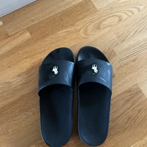 Polo Ralph Lauren tofflor  - Svarta sandaler från Polo Ralph Lauren med klassisk logga i vitt på ovandelen. Tillverkade i syntetmaterial med räfflad sula för extra grepp. Perfekta att slänga på sig till stranden eller när du bara vill chilla.