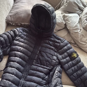 Svart dunjacka från Stone Island - Säljer en svart dunjacka från Stone Island med ikonisk patch på ärmen. Jackan har huva, dragkedja framtill och en bröstficka. Tillverkad i lätt nylon och fylld med dun för extra värme. Perfekt för kalla dagar och har en clean, sportig look. Priset är för att det finns lite hål mm, priset kan även diskuteras! Kom gärna med fler frågor och så kan du få fler bilder privat
