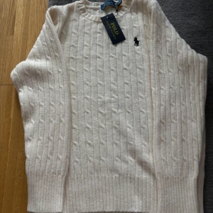 Vit kabelstickad tröja Polo Ralph Lauren - Vit kabelstickad tröja från Polo Ralph Lauren med klassisk rund halsringning och broderad svart logga på bröstet. Tröjan är i mjuk ull och har ribbade muddar vid ärmslut och nederkant. Perfekt för en stilren och tidlös look.