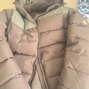 Svart och silver Moncler dunjacka - Säljer en snygg svart dunjacka från Moncler med coola silvriga detaljer över axlarna. Jackan har en vadderad pufferstil, huva och dragkedja framtill. Perfekt för kalla dagar när du vill hålla stilen och värmen.