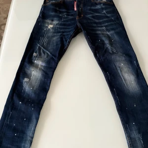 Dsquared2 blå jeans med slitningar - Snygga blå jeans från Dsquared2 med coola slitningar, färgstänk och unika detaljer. Jeansen har klassisk femficksdesign, orange kontrastsömmar och Dsquared2-logga bak. Perfekt för dig som gillar streetstyle och vill sticka ut lite extra.