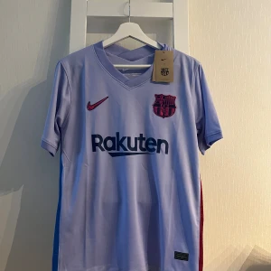 Barça bortatröja Nike M ljuslila - Säljer en FC Barcelona bortatröja från Nike i storlek M. Tröjan är ljuslila med klubbmärke, Rakuten-tryck och La Liga-patch på ärmen. Tillverkad i Dri-FIT-material som andas, perfekt för match eller träning. Klassisk V-ringning och korta ärmar.