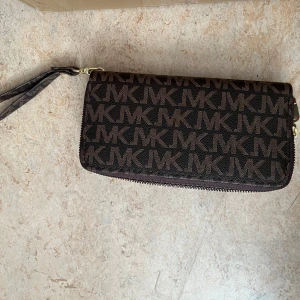 Brun MK plånbok med logomönster - Snygg brun plånbok från Michael Kors med MK-logga över hela ytan. Plånboken har dubbla dragkedjor i guld, flera kortfack och handledsrem. Tillverkad i tåligt syntetmaterial och har ett stilrent, rektangulärt format.