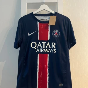 PSG Dembele matchtröja Nike S - Paris Saint-Germain fotbollströja med O. Dembélé och nummer 10 på ryggen. Mörkblå med röd och vit rand på framsidan, PSG-emblem och Qatar Airways-tryck. Tillverkad i lätt och ventilerande material, perfekt för fotbollsplanen.