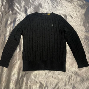  Ralph Lauren tröja  - Kabelstickad svart tröja från Polo Ralph Lauren med klassisk rund halsringning och broderad logga på bröstet. Långärmad modell i mjuk ullblandning, perfekt för lager-på-lager. Snygg ribbad kant vid ärmslut och nederkant.
