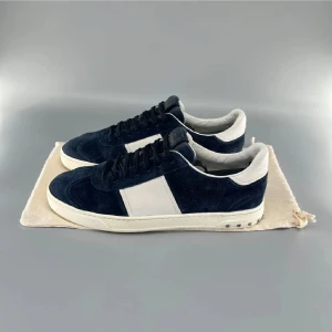 Valentino Garavani sneakers mocka blå/vit - Snygga sneakers från Valentino Garavani i mörkblå mocka med vita detaljer och platt sula. Skorna har svarta skosnören, vita partier på sidan och häl, samt coola nitar baktill på sulan. Yttersulan har ett unikt camouflage-mönster. Perfekta för dig som gillar exklusiva och trendiga sneakers.