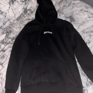 Svart Palm Angels hoodie med tryck - Svart hoodie från Palm Angels i 100% bomull med stor vit logga på ryggen och mindre logga på bröstet. Klassisk känguruficka, snörning i huvan och ribbade muddar. Perfekt för dig som gillar streetwear och vill sticka ut med en ikonisk look. Har en litet hål i armen 