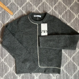 Grå stickad kofta från Zara S - Mysig grå stickad tröja från Zara i storlek S. Koftan har ett unikt vitt kantband som går längs framsidan och bak, rund hals och långa ärmar. Perfekt för dig som gillar stilrena detaljer och vill ha något som känns både chill och snyggt.