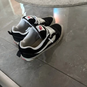 Svarta och vita Vans sneakers - Klassiska Vans sneakers i svart och vitt med vit sula och snörning. Skorna har den ikoniska sidoremsan och Vans-logga på plösen. Ovandelen är i mocka och textil, med rund tå och platt sula. Perfekta för en avslappnad streetstil.