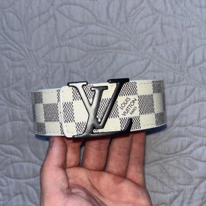 L V bälte - Säljer ett créme färgat bälte från L V med klassiskt rutigt mönster i vitt och grått. Bältet har en stor metallspänne med L V-loggan